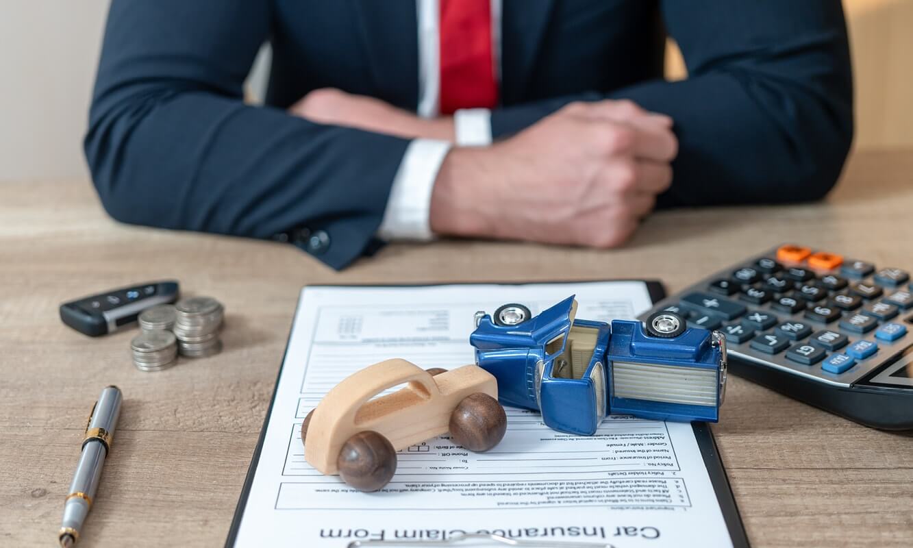 Two toy cars simulate a car accident, on top of an insurance claim form, with coins and a calculator on the desk beside it. A suited figure leans forward on the desk with crossed arms.